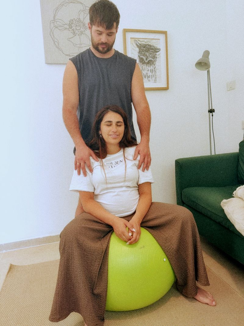 אישה בהריון יושבת על כדור פיזיו ובן זוגה מניח ידיים על כתפיה